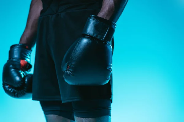 Vista parcial del deportista afroamericano en pantalones cortos y guantes de boxeo sobre fondo azul - foto de stock