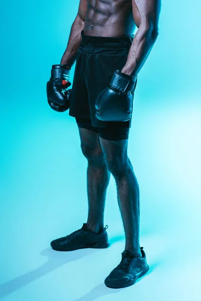 Partial view of african american sportsman in shorts and boxing gloves on blue background — Stock Photo