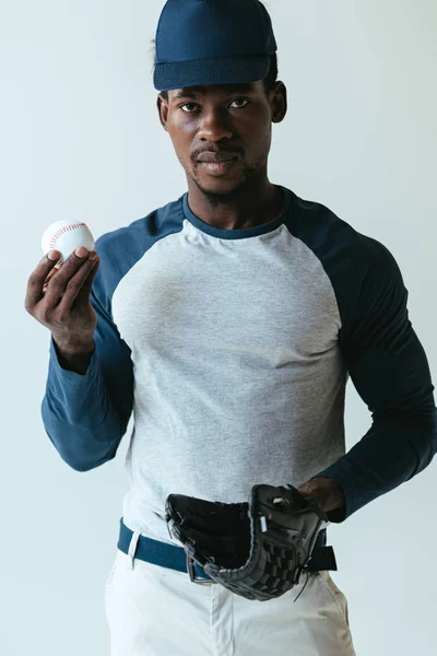 Confiado jugador de béisbol afroamericano mirando a la cámara mientras sostiene la pelota aislada en gris - foto de stock