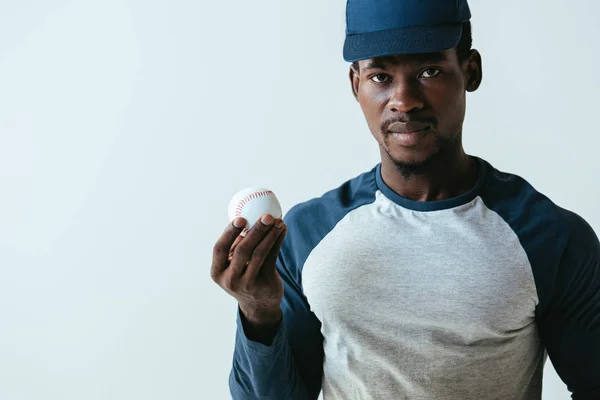 Schöner afrikanisch-amerikanischer Baseballspieler schaut in die Kamera, während er Ball isoliert auf grau hält — Stockfoto