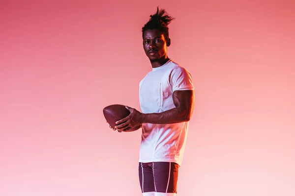 Уверенный в себе американский спортсмен держит регбийный мяч на розовом фоне с градиентом — стоковое фото