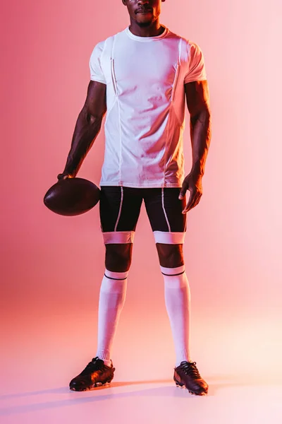 Обрезанный вид африканского американского спортсмена, держащего регбийный мяч на розовом фоне с градиентом и освещением — стоковое фото