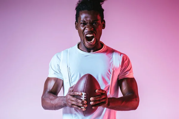 Возбужденный африканский американский спортсмен кричит на камеру, держа регбийный мяч на фиолетовом фоне с градиентом — стоковое фото