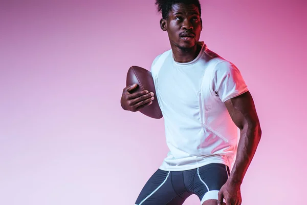 Apuesto afroamericano deportista jugando fútbol americano sobre fondo púrpura con gradiente - foto de stock