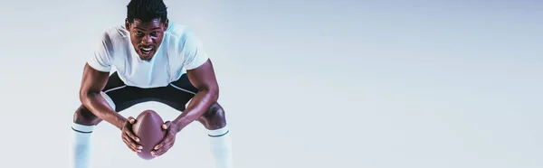 Plano panorámico del deportista afroamericano gritando mientras sostiene la pelota de rugby sobre fondo púrpura con gradiente - foto de stock