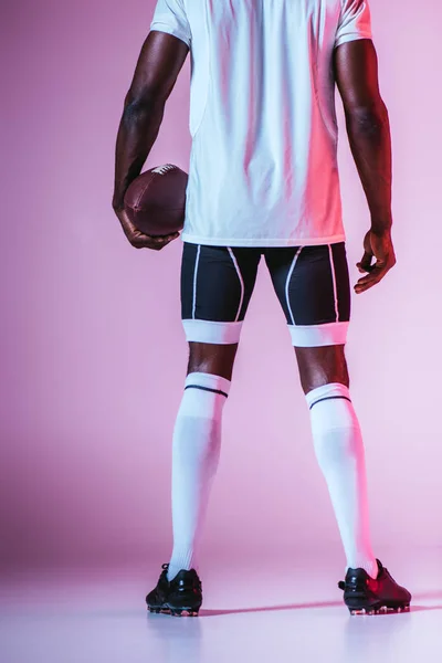 Назад вид африканского американского спортсмена, держащего регбийный мяч на фиолетовом фоне с градиентом и освещением — стоковое фото
