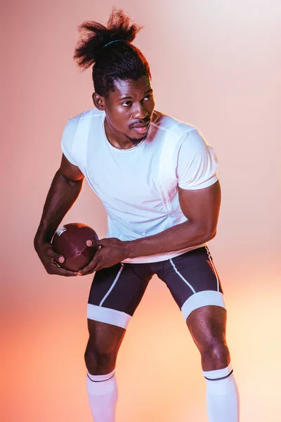 Fiducioso sportivo afroamericano che gioca a calcio americano su sfondo rosa con sfumatura e illuminazione gialla — Foto stock