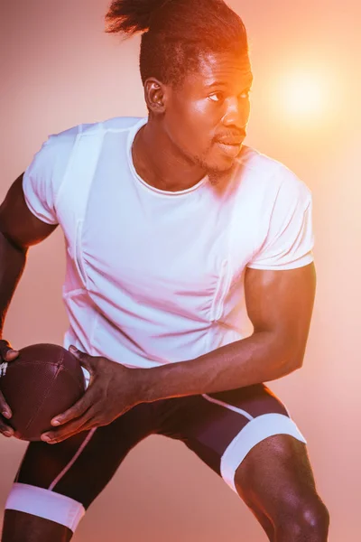 Apuesto deportista afroamericano jugando fútbol americano sobre fondo rosa con gradiente e iluminación amarilla - foto de stock