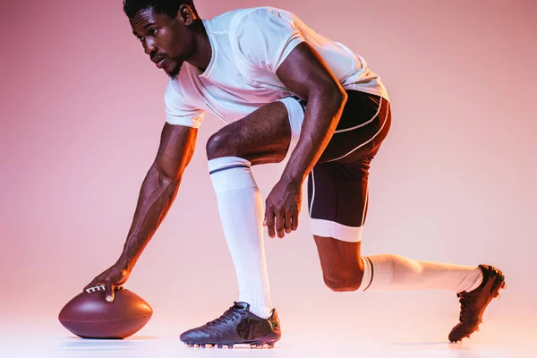 Jovem esportista afro-americano jogando futebol americano em fundo rosa com gradiente e iluminação amarela — Fotografia de Stock