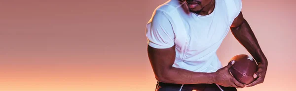 Vista recortada del deportista afroamericano jugando fútbol americano sobre fondo rosa oscuro con gradiente e iluminación, tiro panorámico - foto de stock