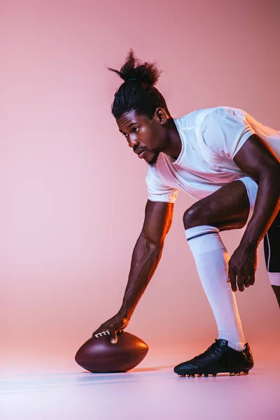 Jovem esportista afro-americano jogando futebol americano em fundo rosa com gradiente e iluminação — Fotografia de Stock