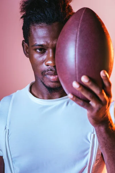 Serious african american sportsman looking at camera while holding rugby ball on pink background with lighting — Stock Photo