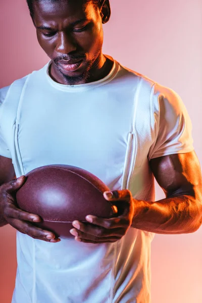 Handsome, young african american sportsman holding rugby ball on pink background with lighting — Stock Photo