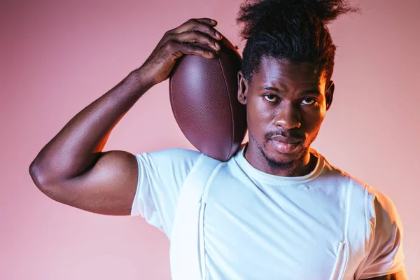 Beau sportif afro-américain regardant la caméra tout en tenant la balle de rugby sur fond rose avec dégradé — Photo de stock