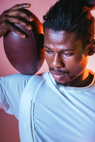 Serio deportista afroamericano sosteniendo pelota de rugby sobre fondo rosa con gradiente e iluminación - foto de stock