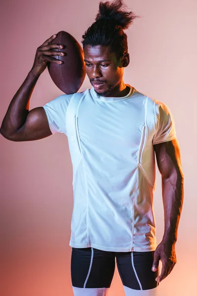 Jovem desportista afro-americano segurando bola de rugby no fundo rosa com gradiente e iluminação — Fotografia de Stock