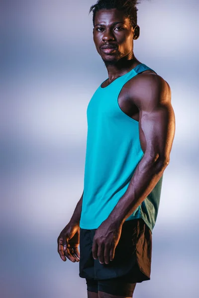 Bello, atletico sportivo afroamericano guardando la fotocamera su sfondo grigio e blu con illuminazione — Foto stock