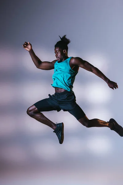 Joven deportista afroamericano atlético saltando sobre fondo gris con iluminación - foto de stock