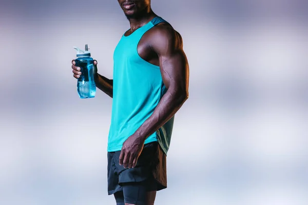 Vista recortada del deportista afroamericano musculoso sosteniendo botella de deportes sobre fondo gris con iluminación - foto de stock