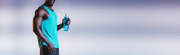 Cropped view of african american sportsman holding sports bottle on grey background with lighting, panoramic shot — Stock Photo