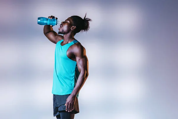 Молодой африканский американский спортсмен, пьющий из спортивной бутылки на сером фоне с освещением — стоковое фото