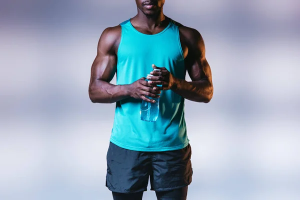 Vue partielle d'un sportif afro-américain musclé tenant une bouteille de sport sur fond gris avec éclairage — Photo de stock