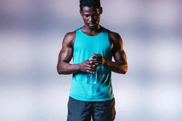 Sportif afro-américain sérieux tenant bouteille de sport sur fond gris avec éclairage — Photo de stock