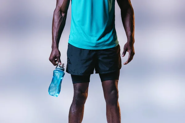 Vista cortada do esportista afro-americano em shorts segurando garrafa de esportes em fundo cinza com iluminação — Fotografia de Stock