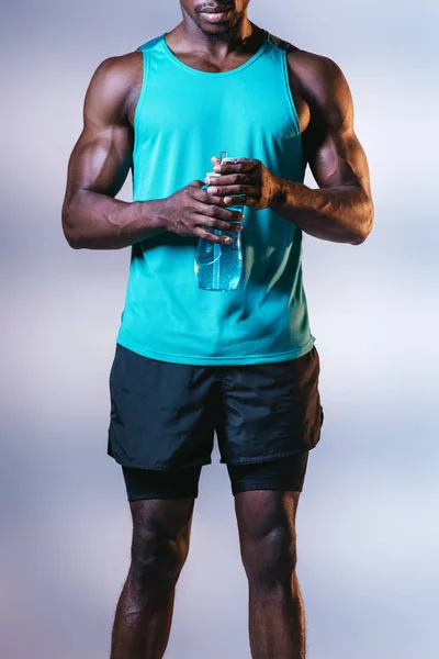 Частичный вид африканского американского спортсмена в шортах, держащего спортивную бутылку на сером фоне с освещением — стоковое фото