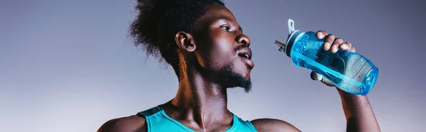 Plano panorámico del deportista afroamericano bebiendo de la botella de deportes sobre fondo gris y azul degradado con iluminación - foto de stock