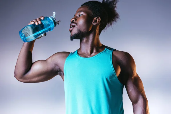 Giovane, muscoloso sportivo afroamericano che beve dalla bottiglia sportiva su sfondo grigio e blu sfumato con illuminazione — Foto stock