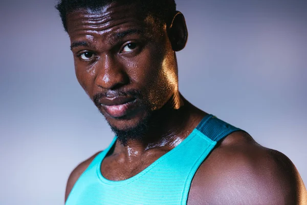 Apuesto, confiado deportista afroamericano con la cara sudorosa mirando a la cámara en gris y azul gradiente fondo con iluminación - foto de stock