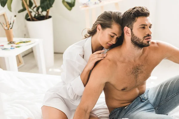 Young sexy couple sitting in bed and hugging while girl sitting with closed eyes and man looking away — Stock Photo