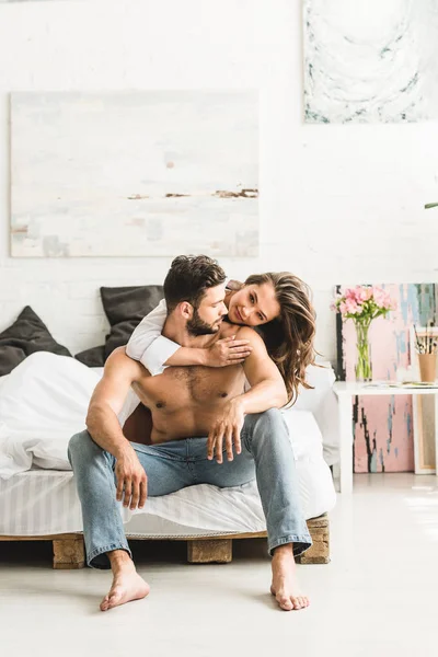 Jovem casal sentado na cama, abraçando e olhando um para o outro — Fotografia de Stock