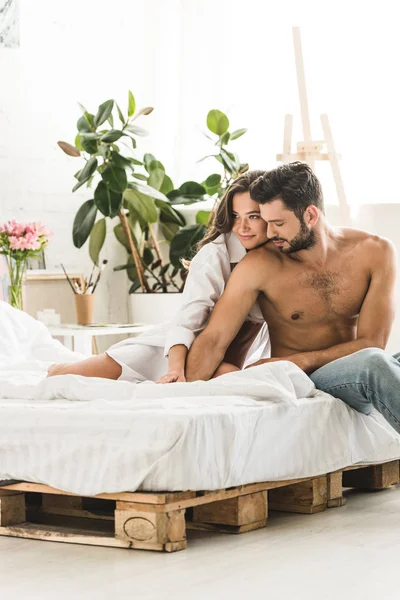 Junges Künstlerpaar, das im Bett sitzt, sich umarmt und Händchen hält — Stockfoto