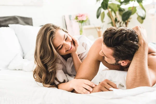 Joven pareja sonriendo mientras yacía en la cama y mirándose - foto de stock