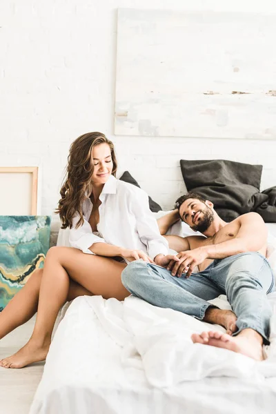 Joven chica en camisa blanca sentado en la cama mientras el hombre acostado en la cama y mirando a la novia - foto de stock