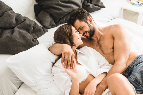 Sexy joven pareja acostada en la cama, sonriendo y abrazando mientras toma de la mano - foto de stock