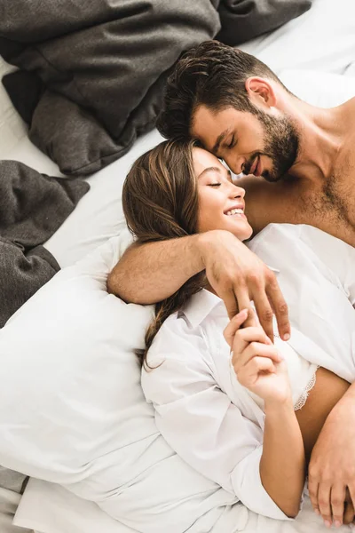 Sexy joven pareja acostada en la cama, sonriendo con los ojos cerrados, abrazando y tomándose de la mano - foto de stock