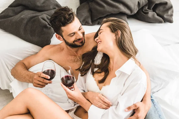 Sexy young couple sitting in bed and smiling while drinking wine — Stock Photo