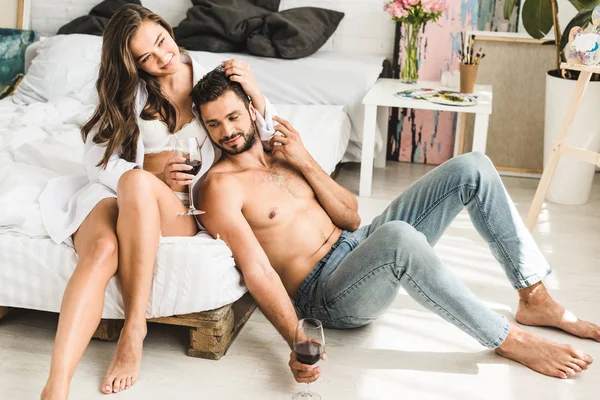 Jeune fille assise dans le lit et touchant les cheveux de petit ami tandis que l'homme assis près du lit et tenant un verre avec du vin — Photo de stock