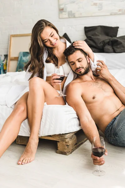 Chica joven sentada en la cama, sonriendo y tocando el pelo del novio mientras el hombre sentado cerca de la cama y sosteniendo el vaso con vino - foto de stock