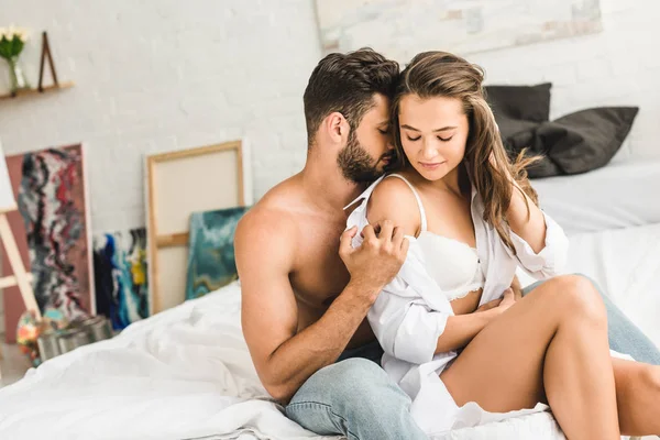 Sexy couple sitting in bed while man trying to put off shirt from girl — Stock Photo
