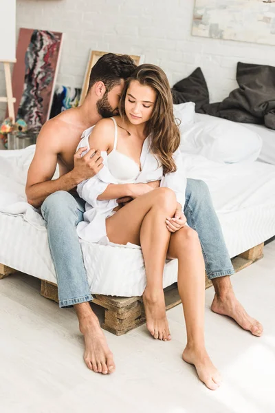 Вид в полный рост сексуальной пары, сидящей в постели, пока мужчина пытается снять рубашку с девушки — стоковое фото