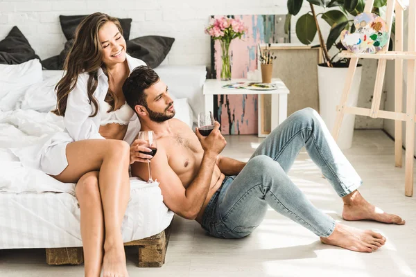 Chica joven sentada en la cama, riendo y tocando el pelo del novio mientras el hombre sentado cerca de la cama y sosteniendo el vaso con vino — Stock Photo