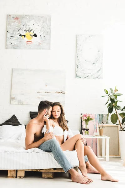 Sexy jovem casal sentado na cama enquanto homem abraçando menina de volta — Fotografia de Stock