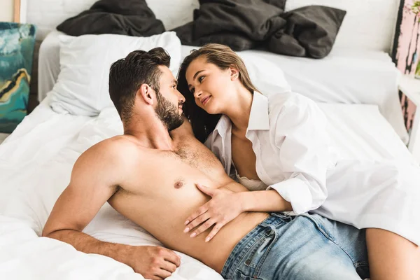 Reizvolles junges Paar liegt im Bett, während Mädchen Oberkörper des Mannes berührt und Freund anschaut — Stockfoto