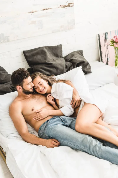 Sexy joven pareja acostado en la cama y sonriendo mientras chica tocando sin camisa hombre - foto de stock