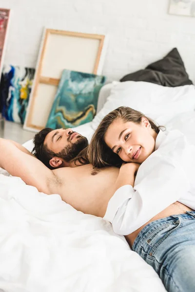 Девушка лежит на мужчине без рубашки и смотрит в камеру, пока парень спит — стоковое фото