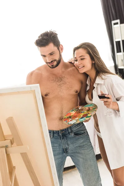 Сексуальный мужчина рисует в то время как девушка стоит рядом с парнем, улыбаясь и держа стакан с вином — стоковое фото